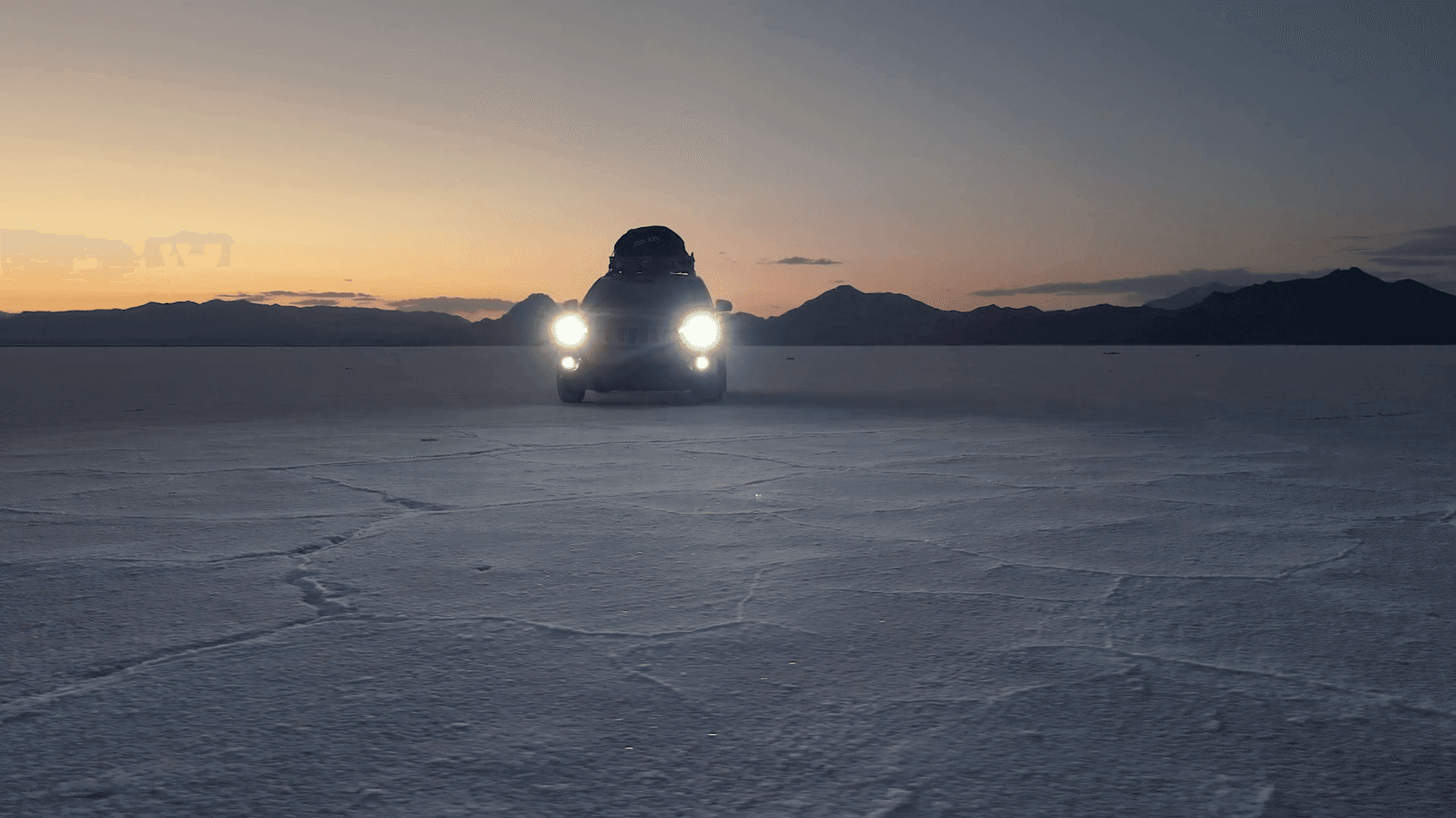 SUV with headlights on, parked on a salt flat at sunset